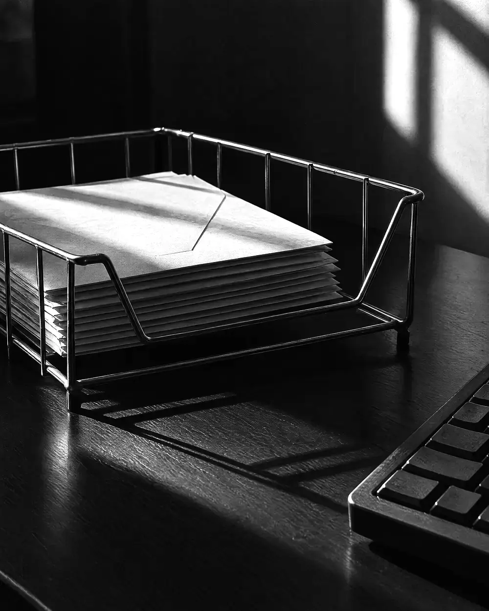 Draht-Ablagekorb mit einem Stapel Briefe neben der Ecke einer Tastatur auf einem dunklen Schreibtisch, tiefe Schatten