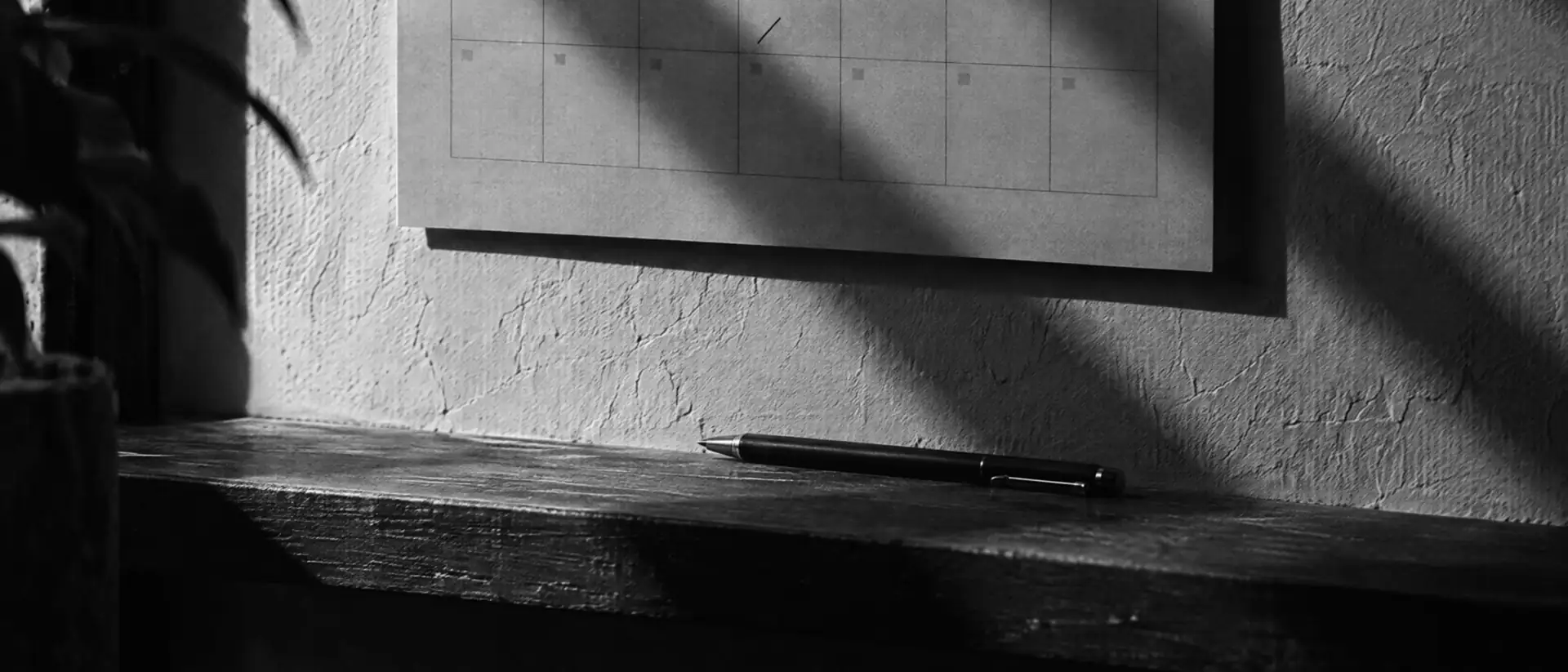 Wandkalender mit leerem Raster an einer Wand, darunter ein Stift auf einer Holzablage im harten Seitenlicht